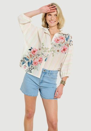 Femme souriante, main levée sur le front, portant un chemisier rayé fleuri et un short en jean bleu clair sur fond uni.