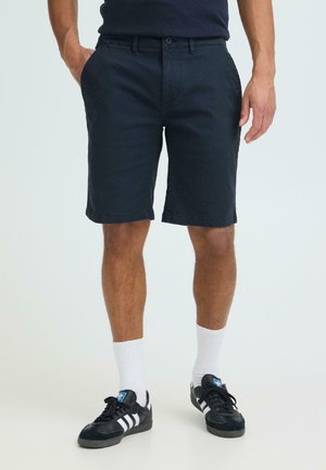 Homme portant un short bleu marine foncé, des chaussettes blanches montantes et des baskets Adidas noires à rayures blanches, debout les mains dans les poches.