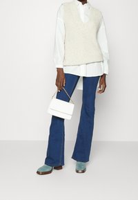 Beige stickad väst över en vit skjorta med knappar, blå utsvängda jeans och gröna mocka anklarstövlar, håller en liten vit handväska med kedja.