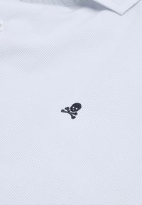 Witte poloshirt met een gestructureerd vierkant patroon, voorzien van een klein zwart doodskop en kruisbotten logo aan de linkerkant. Drieknoopsluiting.