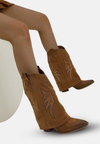 Botas de vaquero de ante marrón con puntas afiladas, que presentan diseños de llamas bordados en un tono más oscuro, y tacones apilados.