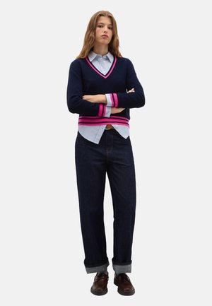 Marineblauer V-Ausschnitt-Pullover mit pinken Streifen, über einem blau-weiß gestreiften Hemd getragen, kombiniert mit dunklen Jeans und braunen Schuhen.