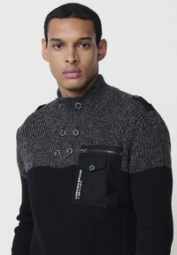 Suéter de punto para hombre en negro y gris con cuello alto, textura acanalada, panel de cuerpo en contraste, botones ornamentales y un bolsillo con cremallera.