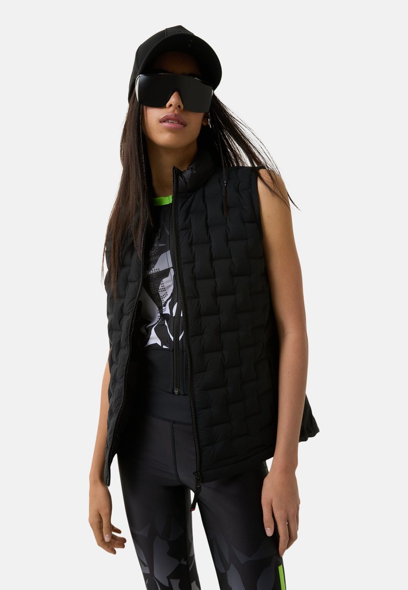 Schwarze gesteppte Weste mit hohem Kragen, ärmellosem Design, kombiniert mit einem grafischen Oberteil und schwarzen Leggings mit einem geometrischen Muster.