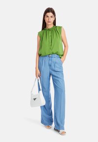 Grünes ärmelloses Oberteil mit gerafftem Ausschnitt, kombiniert mit hellblauen weit geschnittenen Hosen. Die Accessoires umfassen eine weiße Handtasche und offene Schuhe.