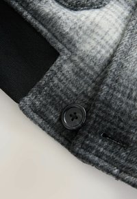 Grå og sort uldblandingsjakke med en struktureret overflade og en sort knap som lukningsdetalje på forsiden.