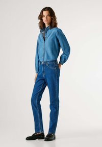 Džínová košile s knoflíkovým zapínáním, spárovaná s džínami vysokého pasu v modré barvě. Černé boty dotvářejí outfit. Oba kusy oděvu mají standardní střih.