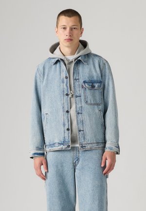 Junger Mann trägt eine hellblaue Jeansjacke und Jeans über einem grauen Hoodie und steht vor einem schlichten Hintergrund.