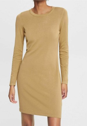 Femme portant une robe en tricot cintrée, à manches longues, couleur camel, avec un col rond, debout devant un fond blanc uni.