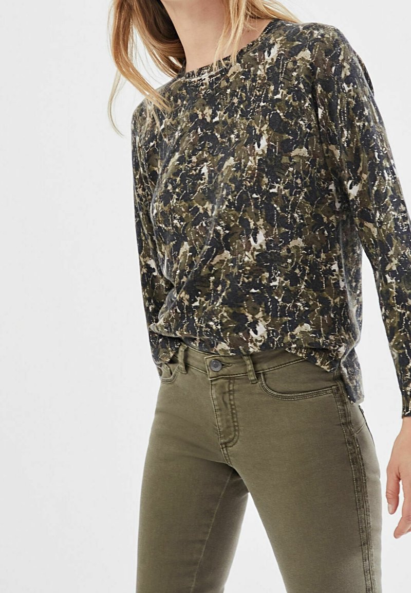 Femme portant un haut à manches longues vert à motif abstrait, rentré dans un jean moulant vert olive, debout devant un fond uni.