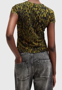 Person som bär en grön och svart leopardmönstrad tajt t-shirt och gråa urblekta jeans med stora bakfickor, handen i fickan.