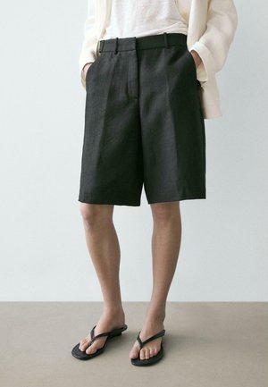 Persona che indossa pantaloncini neri su misura fino al ginocchio, camicia bianca, blazer color crema e sandali infradito neri, in piedi su una superficie uniforme.