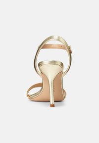 Sandale cu toc înalt metalice aurii, cu o curea îngustă la gleznă și un design cu vârful deschis, având o suprafață netedă și un toc subțire stiletto.