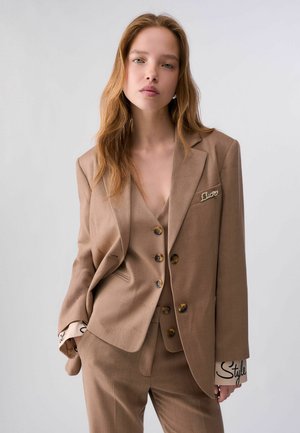 Giovane donna con i capelli lunghi che indossa un completo marrone su misura con gilet e pantaloni, in piedi contro uno sfondo chiaro e uniforme.