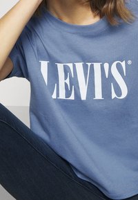 Personne portant un t-shirt Levi's bleu et un jean bleu foncé, avec une partie du visage et du bras visible.