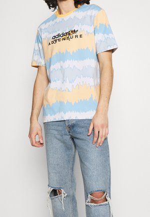 Osoba w niebiesko-żółtej pasiastym t-shircie Adidas Adventure i jasnoniebieskich podartych dżinsach, stojąca na tle jednolitego tła.
