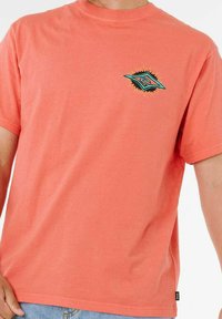 T-shirt di cotone corallo con una piccola grafica in teal e nero sul lato sinistro del petto. Maniche corte e colletto rotondo.