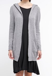 Cardigan grigio a maglia con cappuccio, tasche frontali, apertura frontale e polsini a costine. Indossato sopra un vestito nero, evidenziando la sua versatilità.