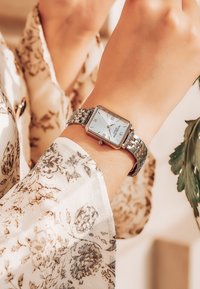 Reloj rectangular plateado con una esfera blanca, números romanos y una pulsera de eslabones exhibido en una muñeca adornada con una manga de patrón floral.