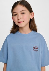 Jasnoniebieska bawełniana koszulka z okrągłym dekoltem i krótkimi rękawami. Koszulka ma małe brązowe logo z tenisówkami na lewej piersi.