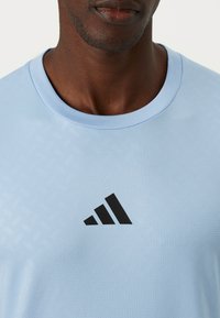 Lys blå sporty t-skjorte med teksturert mønster, rund hals og en svart Adidas-logo på brystet.