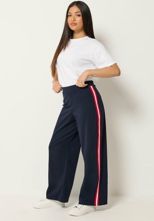 Vrouw draagt een wit T-shirt met korte mouwen en marineblauwe wijde broek met rode en witte zijstrepen, staat met handen in de zij, witte sneakers.