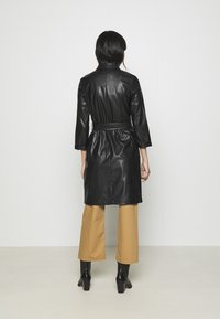 Czarny skórzany płaszcz z paskiem, dopasowanym kołnierzem, długimi rękawami i gładką fakturą, zestawiony z beżowymi szerokimi spodniami i czarnymi botkami na obcasie.