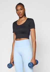 Black ribbed crop top with short sleeves, paired with light blue high-waisted leggings. Model holds blue dumbbells. Plain background.