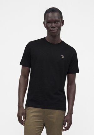 Bărbat purtând un tricou negru cu un mic logo colorat de zebră pe pieptul stâng și pantaloni kaki pe un fundal alb simplu.