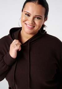 Bruine geribbelde hoodie met een trekkoord in de capuchon. In beeld is een vrouw te zien die de stof bij haar kin vasthoudt, wat de zachte textuur benadrukt.