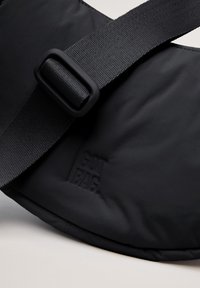 Sort skuldertaske med en tekstureret overflade, der har en bred justerbar rem og et hævet logo-detalje. Slank, buet design.