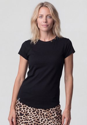 Blonde Frau mit blauen Augen, die ein schwarzes kurzärmeliges Oberteil und einen Rock mit Leopardenmuster trägt, steht vor einem hellgrauen Hintergrund.