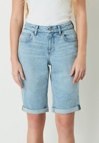 Frau trägt hellblaue Jeansshorts mit umgekrempeltem Saum und ein weißes ärmelloses Top, steht vor einem schlichten hellen Hintergrund.