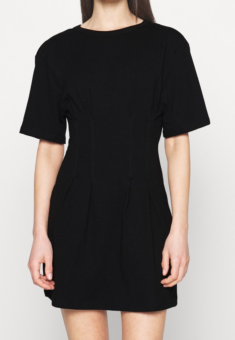 Femme portant une robe noire courte à manches courtes et taille cintrée, visible du cou jusqu'à mi-cuisse sur un fond blanc uni.