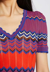 Gros plan d'une femme portant un haut tricoté à motif chevron rouge et bleu, avec un col en V festonné et des manches courtes bouffantes.