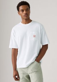 White cotton short-sleeve t-shirt with a chest pocket featuring red embroidered text. Relaxed fit, smooth texture, and round neckline.