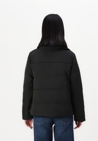 Black puffer jacket with a high collar and rounded shoulders. Features a quilted design and is worn over blue jeans, showing a relaxed fit.