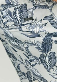 Pantalones cortos de tela gris claro con un estampado de hojas tropicales azules. Cuenta con un bolsillo trasero con una pequeña etiqueta negra.