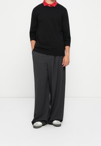 Personne portant un pull noir par-dessus une chemise à col rouge, un pantalon large gris foncé et des baskets grises, debout contre un mur blanc.