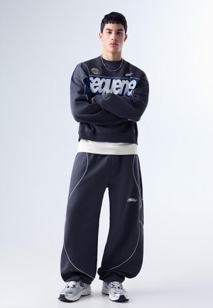 Junger Mann mit verschränkten Armen, der einen schwarz-weißen, übergroßen Trainingsanzug und silberne Sneakers trägt, vor einem weißen Hintergrund.