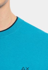 Maglione turchese a maglia con colletto a coste neri e un piccolo dettaglio del logo sul lato sinistro in basso. Texture liscia, design a maniche corte.