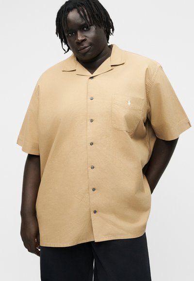 Asmuo su tamsia oda ir dreadykais, dėvintis laisvą smėlio spalvos trumpomis rankovėmis marškinius su krūtinės kišene ir juodas kelnes priešais vientisą foną.