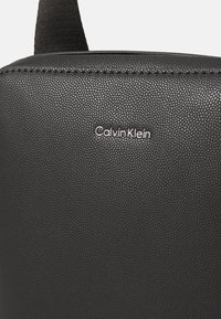 Svart läderväska med struktur och en slät yta, försedd med en liten silverfärgad "Calvin Klein"-logotyp framtill. Minimalistisk design med ett handtag upptill.