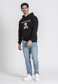 Sudadera negra con un gráfico de oso y el texto "Gianni Kavanagh", combinada con jeans de mezclilla desgastados de color azul claro y zapatillas blancas.