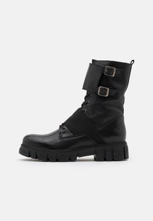 Bota de cuero negro hasta el tobillo con puntera redonda, suela gruesa, dos hebillas laterales y banda elástica. Presenta un acabado mate texturizado y detalles de costura.