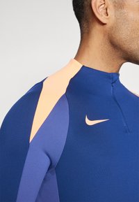 Kék Nike sportfelső barackszínű díszítéssel. Magas gallérral, cipzárral és jellegzetes logóval rendelkezik. Simább szövetből készült.