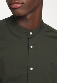 Man in een donkergroene shirt met mandarinekraag, witte knopen en een klein "ck" logo op de linkerkant van de borst, tegen een effen achtergrond.
