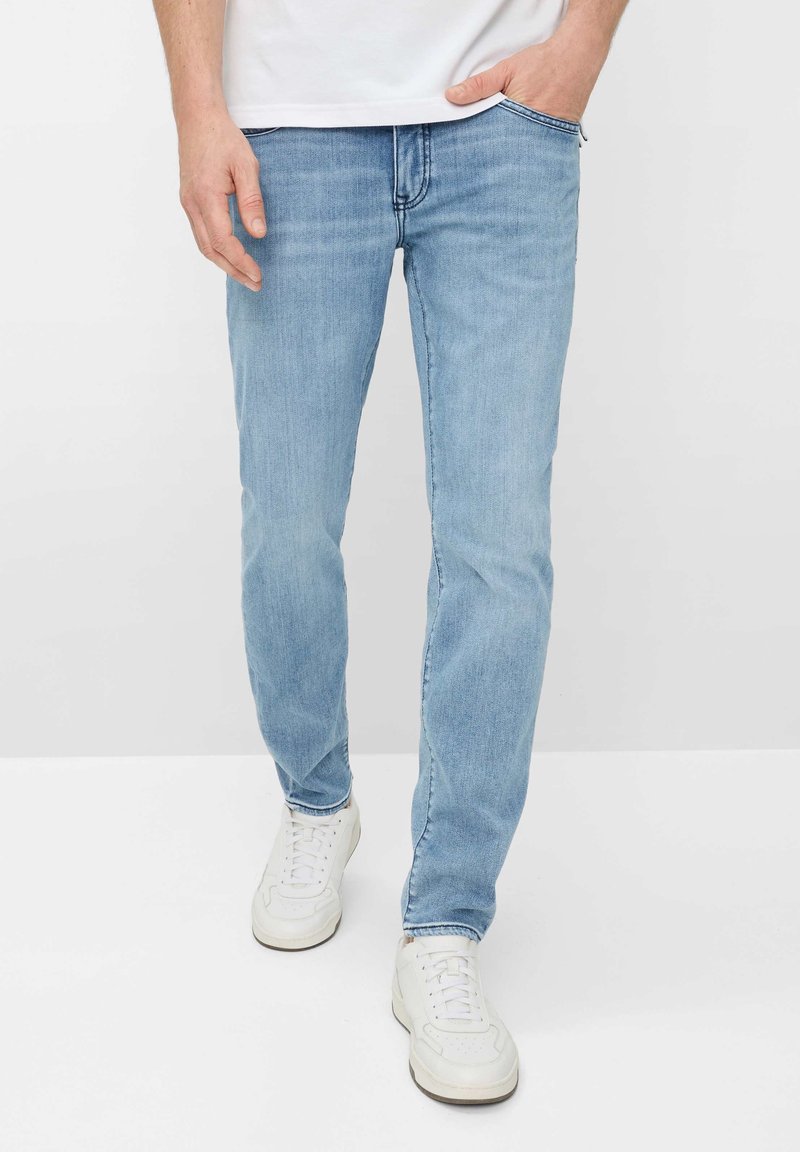 Homme portant un jean droit bleu clair et des baskets blanches, une main dans la poche, debout sur un fond blanc uni.