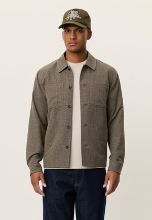 Brown textured button-up jacket with two front pockets, worn over a light-colored shirt, paired with navy jeans and a green cap.