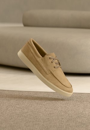 Light brown suede casual lace-up shoe floating above beige carpet with a matching beige sofa blurred in the background.